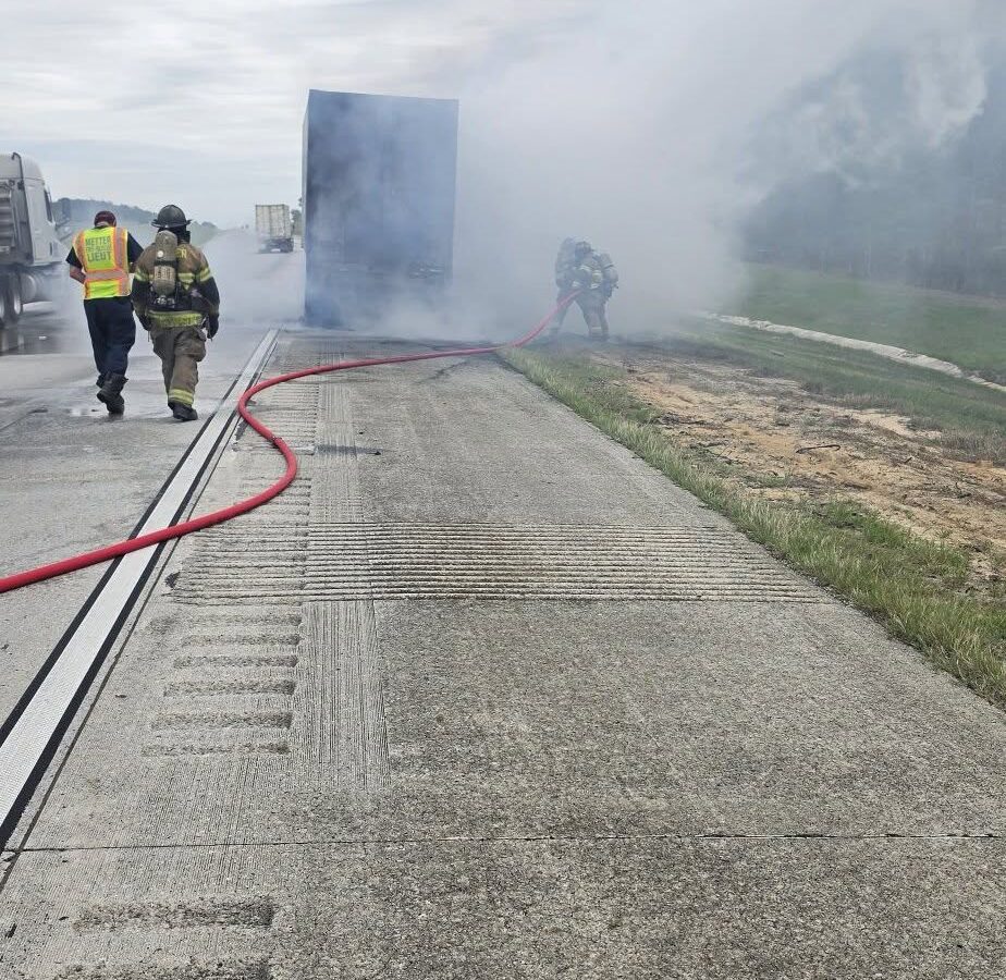 A highway scene with a large truck emitting thick smoke, firefighters actively working to extinguish the fire using a red hose, and another person in a high-visibility vest walking alongside a firefighter. The sky is overcast, and the road surface is textured concrete.