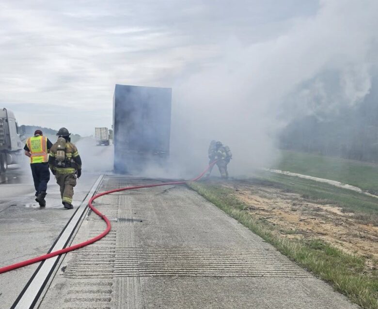 A highway scene with a large truck emitting thick smoke, firefighters actively working to extinguish the fire using a red hose, and another person in a high-visibility vest walking alongside a firefighter. The sky is overcast, and the road surface is textured concrete.