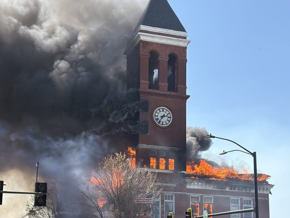 A large brick building with a clock tower is engulfed in flames and thick black smoke. The fire is visible through multiple windows and the roof, with bright orange flames and heavy smoke billowing out. Several people, including emergency responders, are in the foreground, with one holding yellow police tape to cordon off the area. A red fire department vehicle and traffic lights are also visible nearby. The sky is clear and blue.