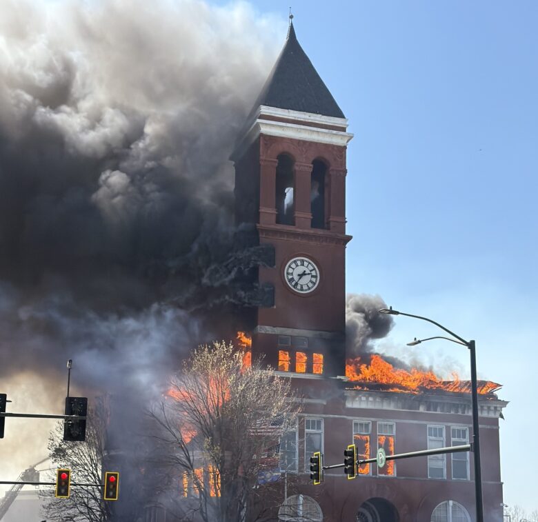 A large brick building with a clock tower is engulfed in flames and thick black smoke. The fire is visible through multiple windows and the roof, with bright orange flames and heavy smoke billowing out. Several people, including emergency responders, are in the foreground, with one holding yellow police tape to cordon off the area. A red fire department vehicle and traffic lights are also visible nearby. The sky is clear and blue.