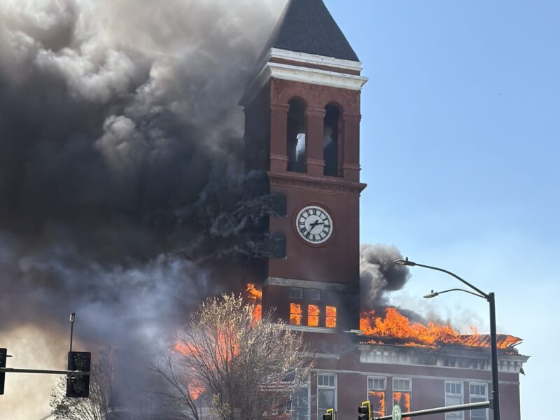 A large brick building with a clock tower is engulfed in flames and thick black smoke. The fire is visible through multiple windows and the roof, with bright orange flames and heavy smoke billowing out. Several people, including emergency responders, are in the foreground, with one holding yellow police tape to cordon off the area. A red fire department vehicle and traffic lights are also visible nearby. The sky is clear and blue.