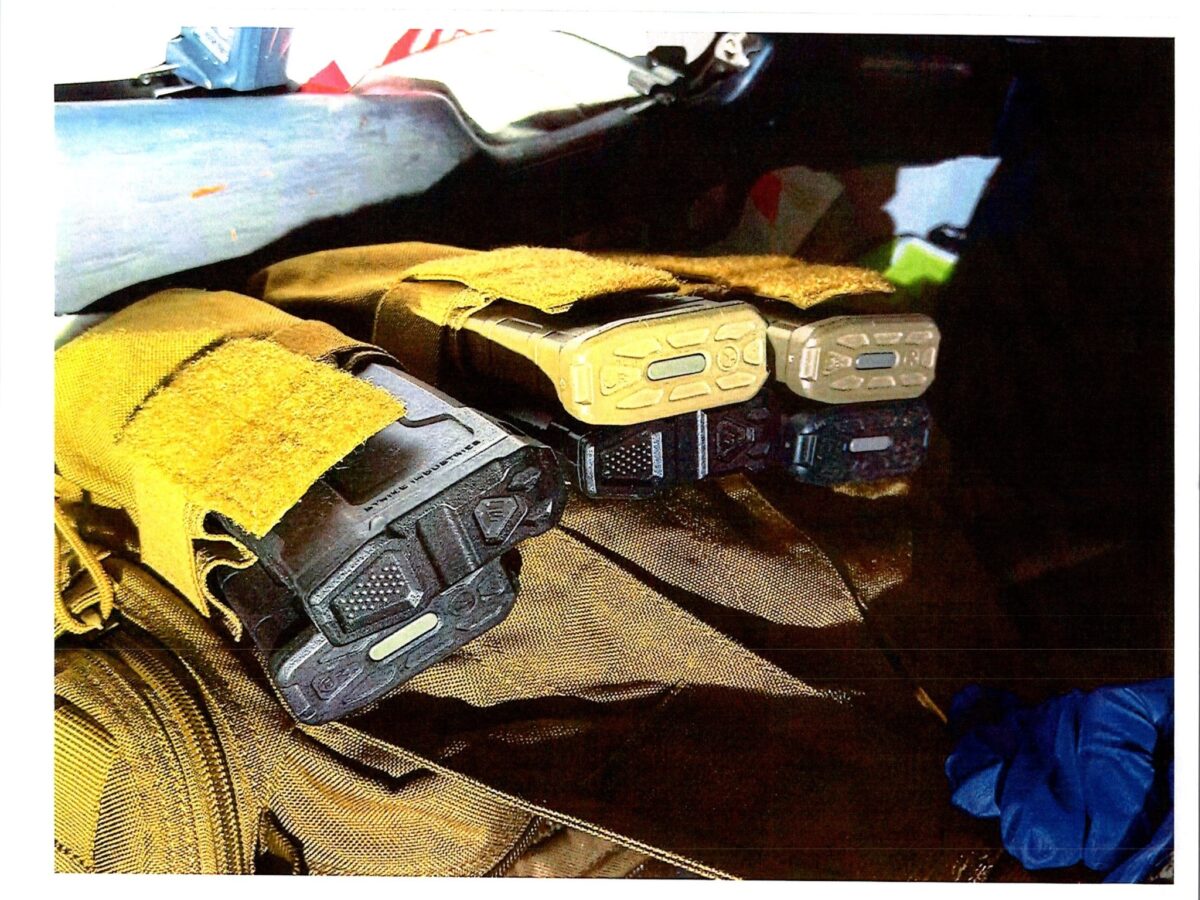 Four rifle magazines are partially covered with yellow fabric sleeves, lying on a brown textured surface. The magazines have visible base plates with different colors and markings. A blue glove is visible in the lower right corner.