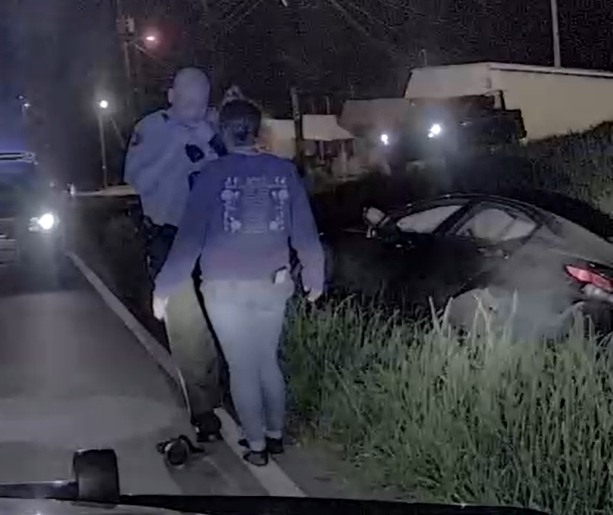 A nighttime scene shows a police officer standing on a road next to a woman wearing a dark hoodie and light jeans. To the right, a dark-colored car is partially off the road, resting in tall grass. The area is dimly lit with some streetlights and vehicle headlights in the background.