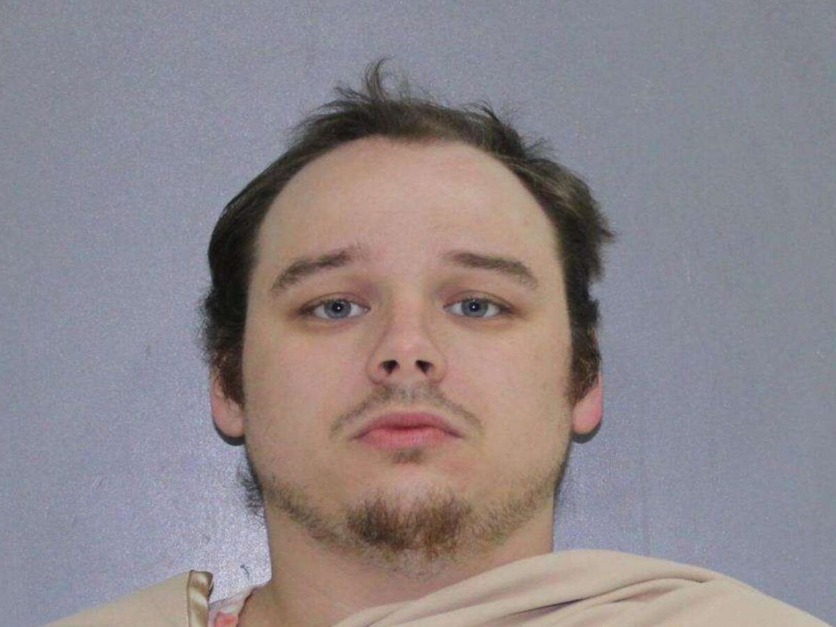 Close-up of a man with light skin, short dark hair, blue eyes, and a patchy beard and mustache, wearing a beige garment against a plain gray background.