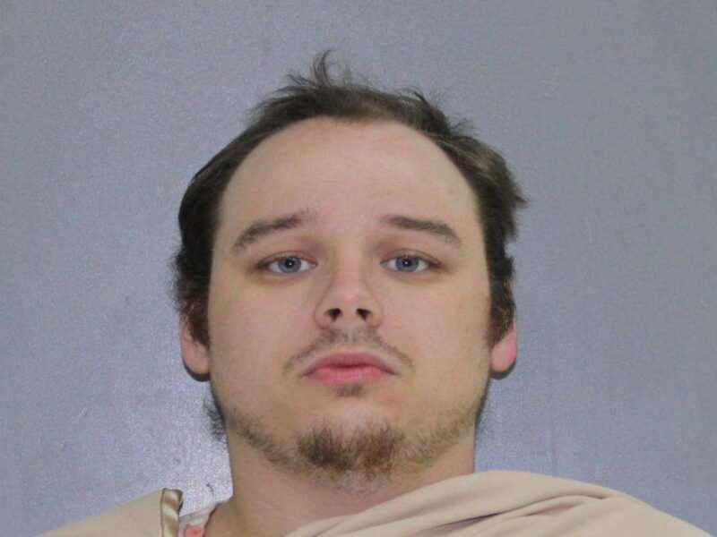 Close-up of a man with light skin, short dark hair, blue eyes, and a patchy beard and mustache, wearing a beige garment against a plain gray background.
