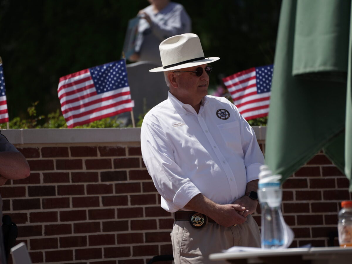 A man wearing a white long-sleeve shirt with a sheriff's badge emblem, beige pants, a white hat with a black band, and sunglasses stands in front of a brick wall. He has a sheriff's badge on his belt and is standing with his hands clasped. Behind him, there are small American flags and a blurred figure. A table with a water bottle and an orange drink is partially visible in the foreground.