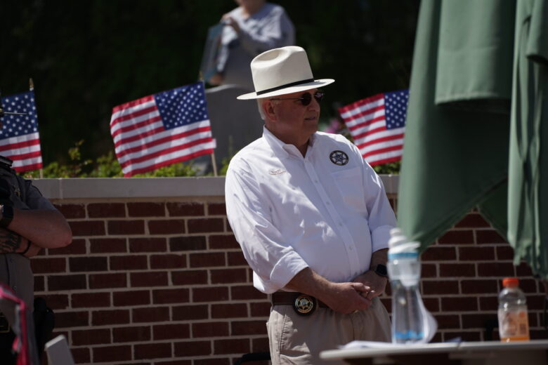 A man wearing a white long-sleeve shirt with a sheriff's badge emblem, beige pants, a white hat with a black band, and sunglasses stands in front of a brick wall. He has a sheriff's badge on his belt and is standing with his hands clasped. Behind him, there are small American flags and a blurred figure. A table with a water bottle and an orange drink is partially visible in the foreground.
