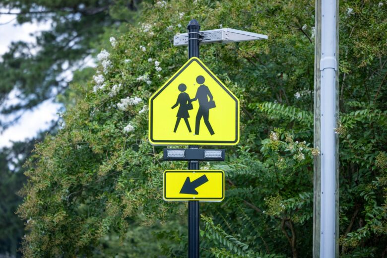 Yellow pedestrian crossing sign with silhouettes of an adult and child holding hands, mounted on a black pole with a solar panel and two rectangular lights above it, and a yellow arrow sign pointing left below it, surrounded by green foliage.