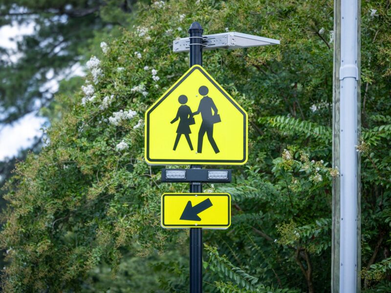 Yellow pedestrian crossing sign with silhouettes of an adult and child holding hands, mounted on a black pole with a solar panel and two rectangular lights above it, and a yellow arrow sign pointing left below it, surrounded by green foliage.