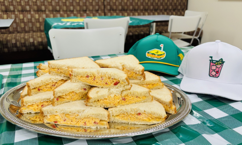 A silver tray holds a stack of triangular pimento cheese sandwiches made with white bread. The tray is placed on a green and white checkered tablecloth. Behind the tray, there are two baseball caps: one green with a sandwich and golf flag logo, and one white with a pink drink and lime logo. The background shows white chairs and a brown patterned bench.