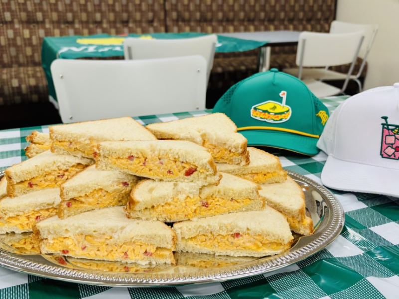 A silver tray holds a stack of triangular pimento cheese sandwiches made with white bread. The tray is placed on a green and white checkered tablecloth. Behind the tray, there are two baseball caps: one green with a sandwich and golf flag logo, and one white with a pink drink and lime logo. The background shows white chairs and a brown patterned bench.
