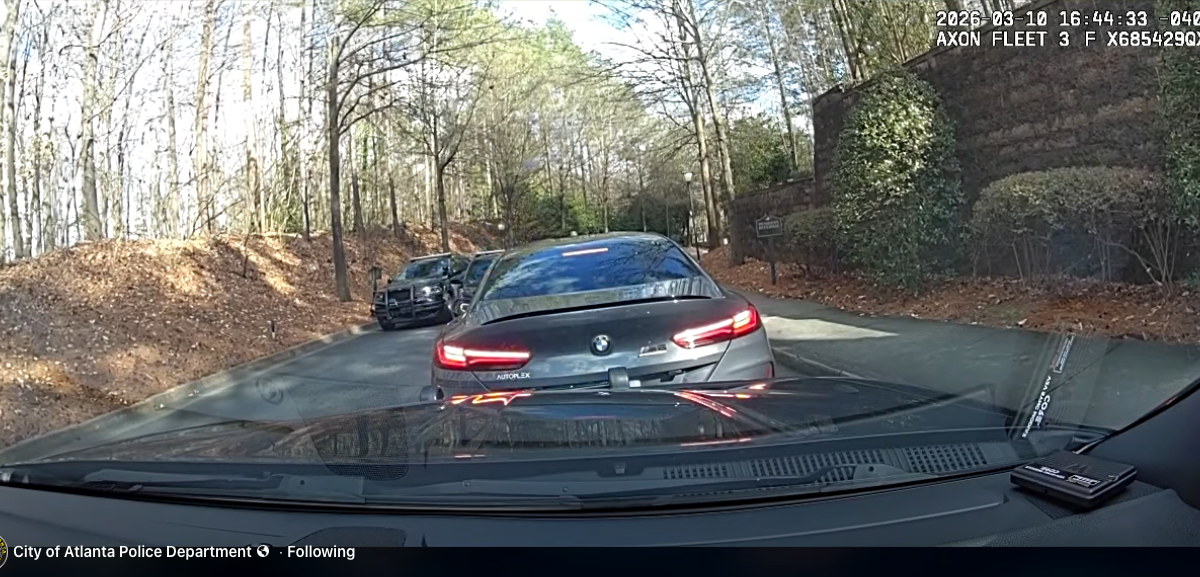View from inside a police vehicle showing a black BMW with its brake lights on stopped on a narrow road lined with trees and bushes. Another police vehicle is visible further ahead on the left side of the road. The timestamp on the image reads "2026-03-10 16:44:33 -0400.