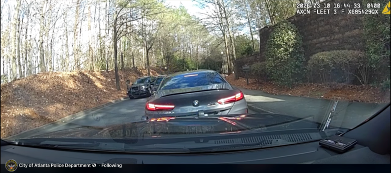 View from inside a police vehicle showing a black BMW with its brake lights on stopped on a narrow road lined with trees and bushes. Another police vehicle is visible further ahead on the left side of the road. The timestamp on the image reads "2026-03-10 16:44:33 -0400.