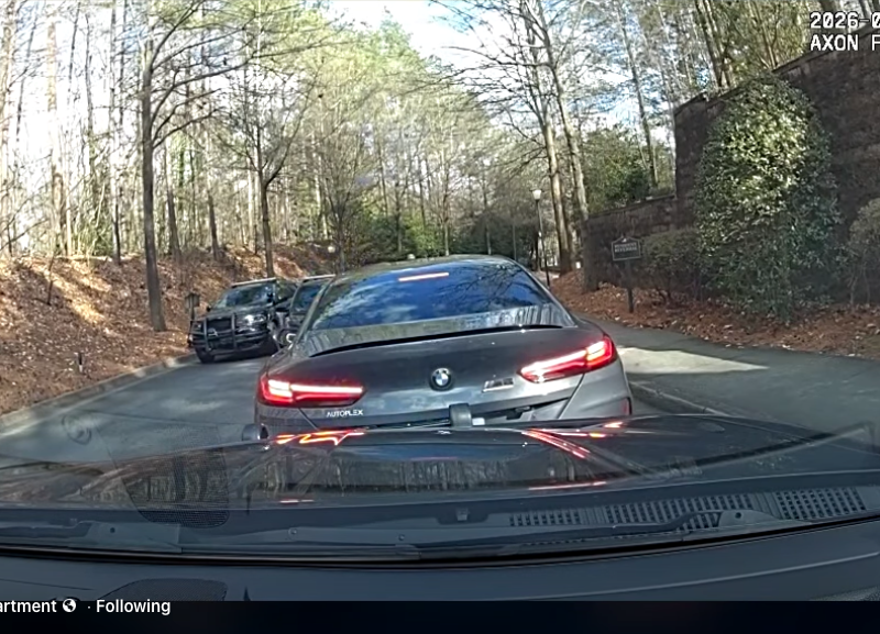 View from inside a police vehicle showing a black BMW with its brake lights on stopped on a narrow road lined with trees and bushes. Another police vehicle is visible further ahead on the left side of the road. The timestamp on the image reads "2026-03-10 16:44:33 -0400.