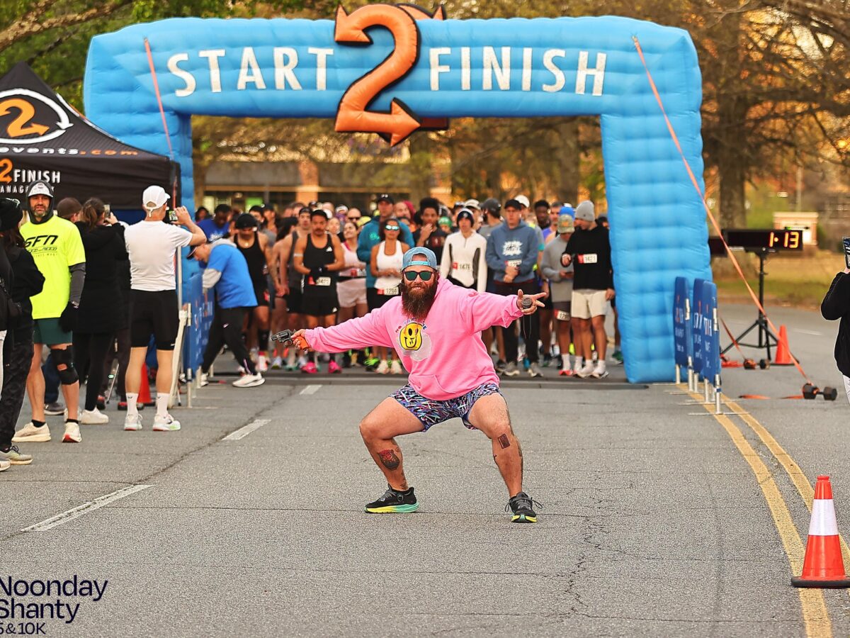 A man in a bright pink hoodie, patterned shorts, sunglasses, and a backward cap is crouching and posing energetically in front of a blue inflatable arch labeled "START 2 FINISH" at a race event. Behind him, a crowd of runners is gathered at the starting line, with some people standing on the sides. The event is identified as the "Noonday Shanty 5 & 10K" in the bottom left corner.