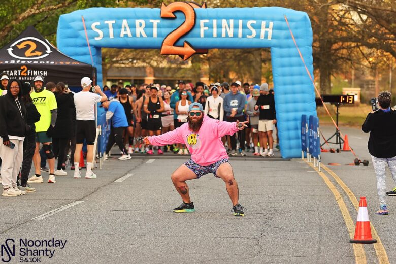 A man in a bright pink hoodie, patterned shorts, sunglasses, and a backward cap is crouching and posing energetically in front of a blue inflatable arch labeled "START 2 FINISH" at a race event. Behind him, a crowd of runners is gathered at the starting line, with some people standing on the sides. The event is identified as the "Noonday Shanty 5 & 10K" in the bottom left corner.