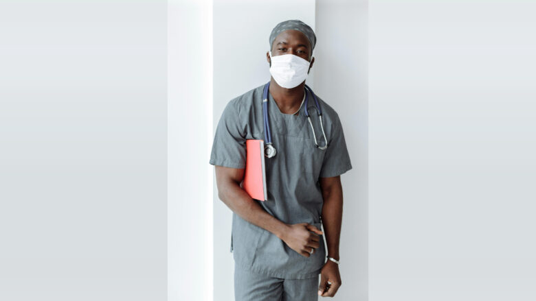 A male healthcare professional wearing gray scrubs, a matching cap, and a white face mask. He has a stethoscope around his neck and is holding a red folder under his left arm. The background is plain white.