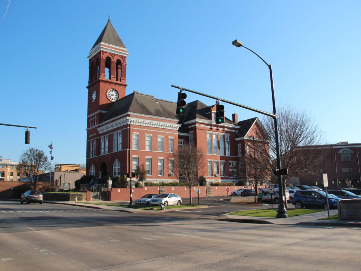 A large red brick building with a tall clock tower and arched windows, situated at a street corner with traffic lights and several parked cars nearby. The sky is clear and blue.
