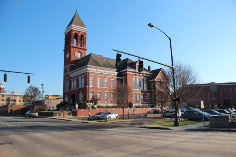 A large red brick building with a tall clock tower and arched windows, situated at a street corner with traffic lights and several parked cars nearby. The sky is clear and blue.