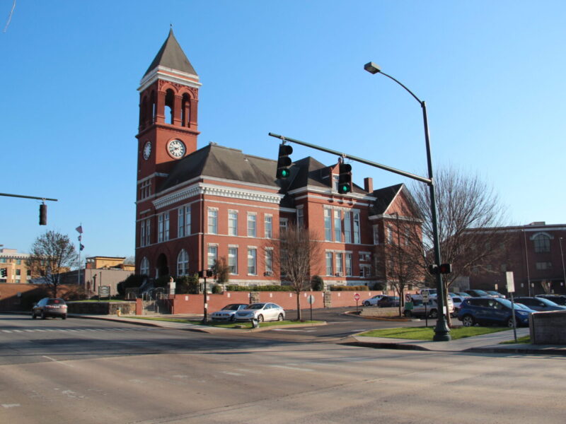 A large red brick building with a tall clock tower and arched windows, situated at a street corner with traffic lights and several parked cars nearby. The sky is clear and blue.