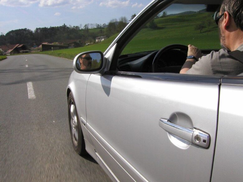 Silver car driving on a rural road with a man wearing sunglasses behind the wheel, visible through the open driver's side window and side mirror. Green fields and houses are in the background under a partly cloudy sky.