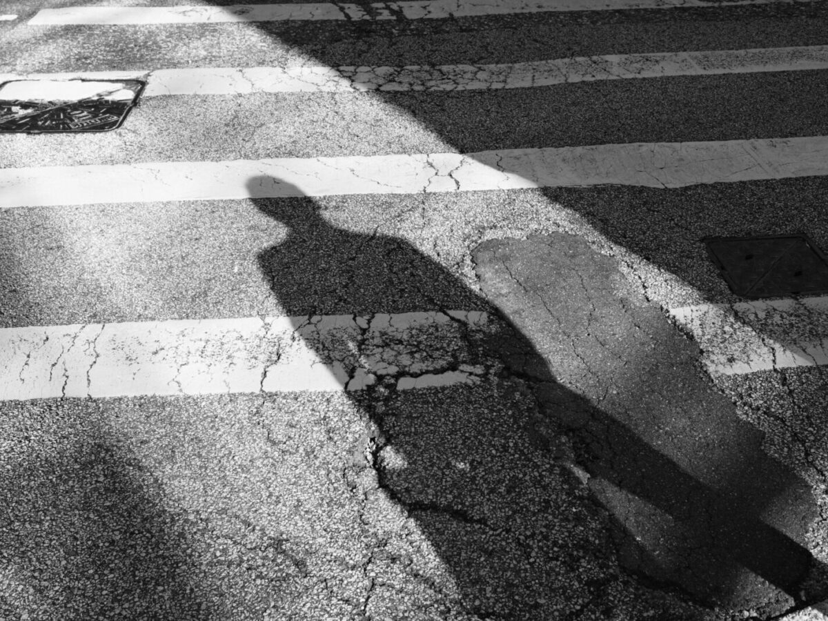 Shadow of a person cast on a cracked asphalt street with white pedestrian crosswalk lines. The image is in black and white, showing texture and contrast between light and shadow.