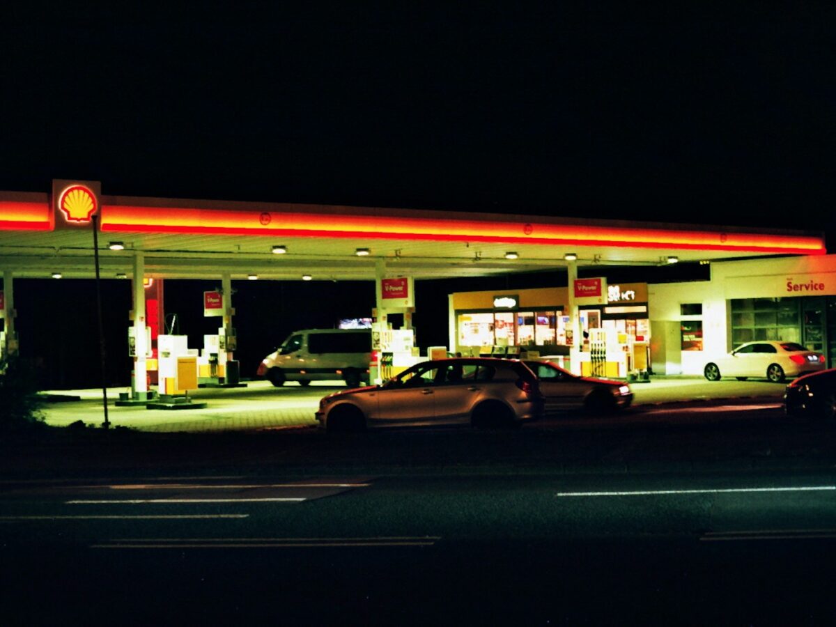 Shell gas station at night with illuminated canopy and fuel pumps. Several cars are parked near the pumps and the convenience store, which is brightly lit. The service area is visible on the right side of the station. The surrounding area is dark.