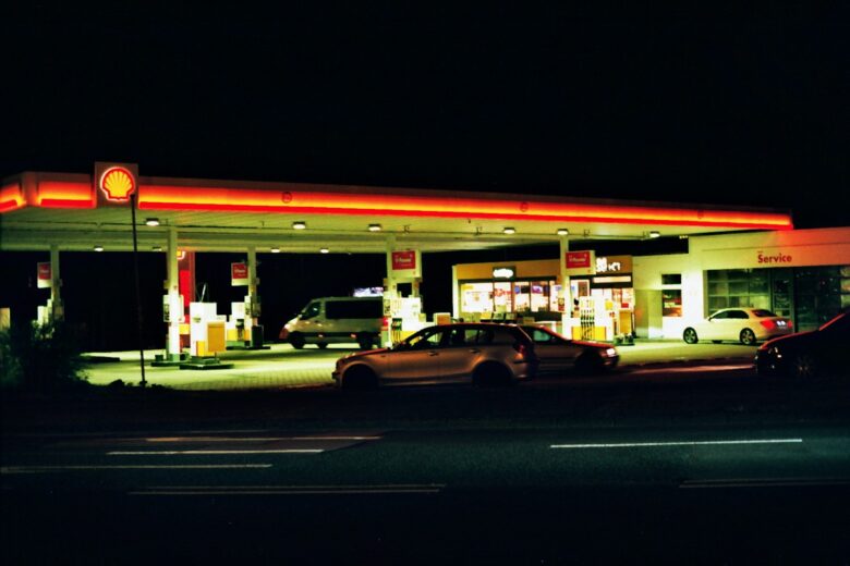 Shell gas station at night with illuminated canopy and fuel pumps. Several cars are parked near the pumps and the convenience store, which is brightly lit. The service area is visible on the right side of the station. The surrounding area is dark.