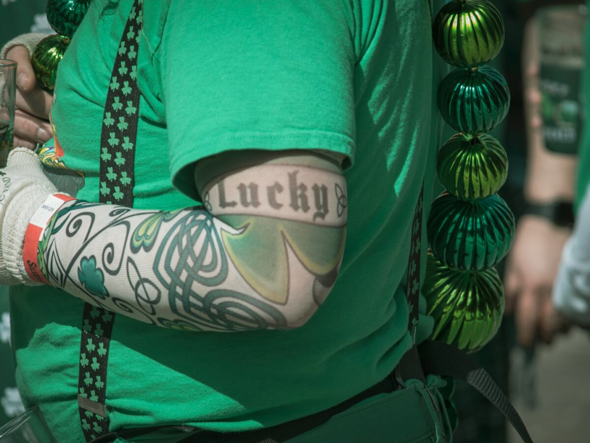 A person wearing a green shirt and shamrock-patterned suspenders has a tattoo sleeve on their arm featuring Celtic designs and a large shamrock. The word "Lucky" is tattooed in black Old English font on the upper arm. The person is also wearing a white glove with green shamrocks and text, holding a drink in a clear plastic cup. Green metallic beads hang from the person's shoulder.