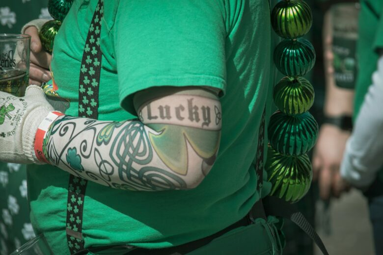 A person wearing a green shirt and shamrock-patterned suspenders has a tattoo sleeve on their arm featuring Celtic designs and a large shamrock. The word "Lucky" is tattooed in black Old English font on the upper arm. The person is also wearing a white glove with green shamrocks and text, holding a drink in a clear plastic cup. Green metallic beads hang from the person's shoulder.