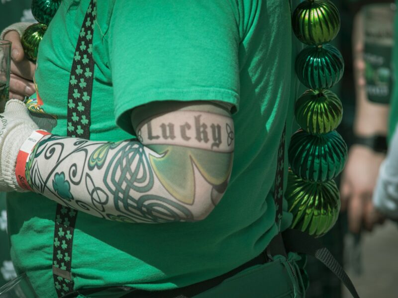A person wearing a green shirt and shamrock-patterned suspenders has a tattoo sleeve on their arm featuring Celtic designs and a large shamrock. The word "Lucky" is tattooed in black Old English font on the upper arm. The person is also wearing a white glove with green shamrocks and text, holding a drink in a clear plastic cup. Green metallic beads hang from the person's shoulder.