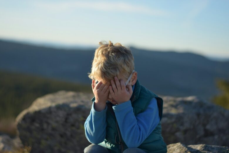 A young boy with blonde hair wearing glasses, a blue long-sleeve shirt, and a green vest is sitting on a rock outdoors with his face covered by both hands. The background shows blurred mountains and a clear sky.