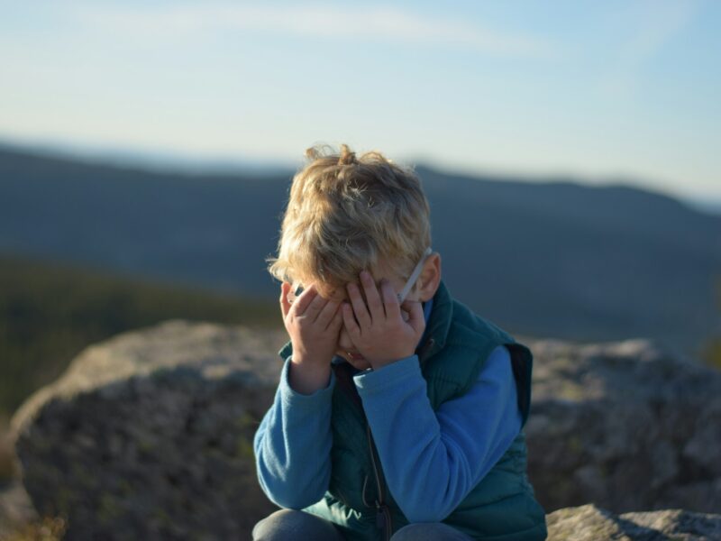 A young boy with blonde hair wearing glasses, a blue long-sleeve shirt, and a green vest is sitting on a rock outdoors with his face covered by both hands. The background shows blurred mountains and a clear sky.