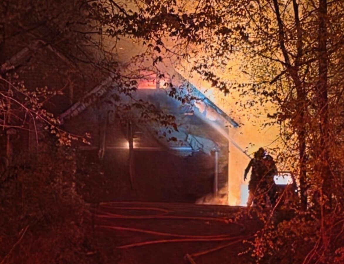 A house engulfed in flames at night, surrounded by trees. Firefighters are actively working to extinguish the fire, with hoses laid out on the ground leading up to the house. The scene is illuminated by the bright orange glow of the fire and red emergency lights.