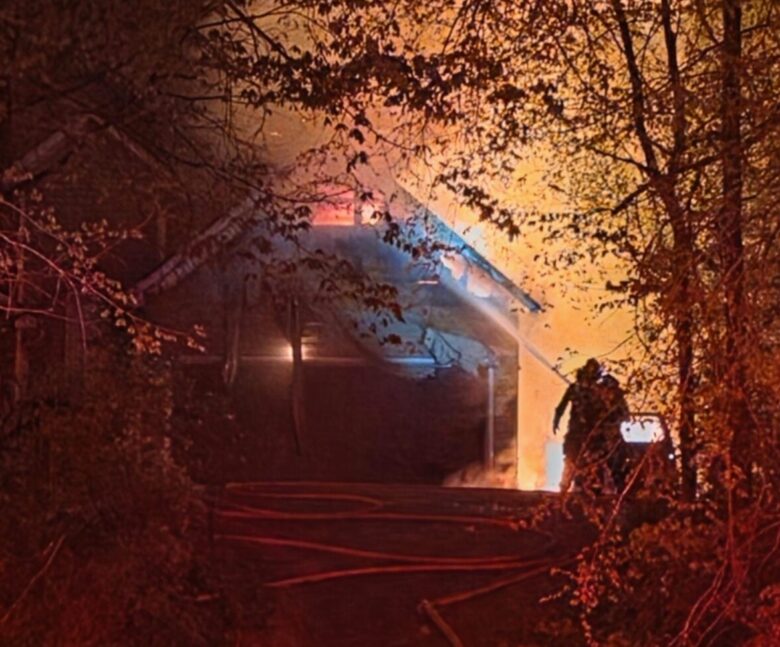 A house engulfed in flames at night, surrounded by trees. Firefighters are actively working to extinguish the fire, with hoses laid out on the ground leading up to the house. The scene is illuminated by the bright orange glow of the fire and red emergency lights.