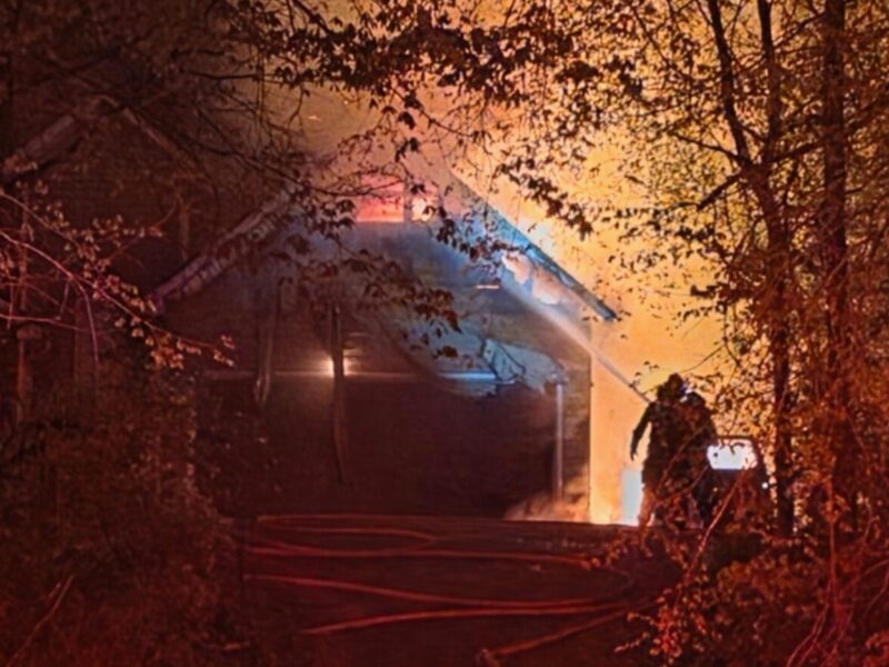 A house engulfed in flames at night, surrounded by trees. Firefighters are actively working to extinguish the fire, with hoses laid out on the ground leading up to the house. The scene is illuminated by the bright orange glow of the fire and red emergency lights.