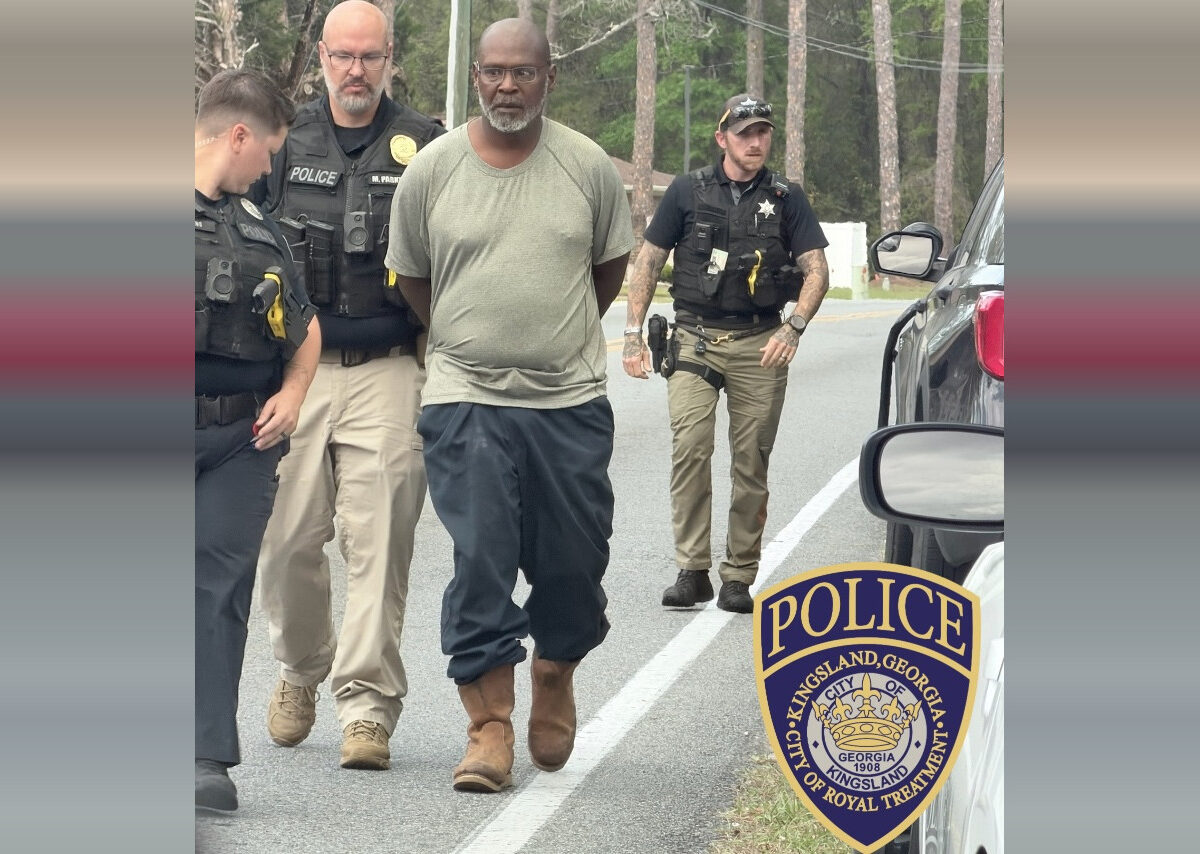 A man with a gray beard and glasses, wearing a light gray t-shirt, dark pants, and brown boots, is being escorted by three police officers on a road. The officers are in uniform with tactical vests, and one officer is walking behind the man while the other two are beside him. The scene takes place outdoors with trees in the background. A police badge for Kingsland, Georgia, is visible in the lower right corner.