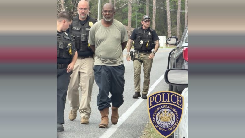 A man with a gray beard and glasses, wearing a light gray t-shirt, dark pants, and brown boots, is being escorted by three police officers on a road. The officers are in uniform with tactical vests, and one officer is walking behind the man while the other two are beside him. The scene takes place outdoors with trees in the background. A police badge for Kingsland, Georgia, is visible in the lower right corner.