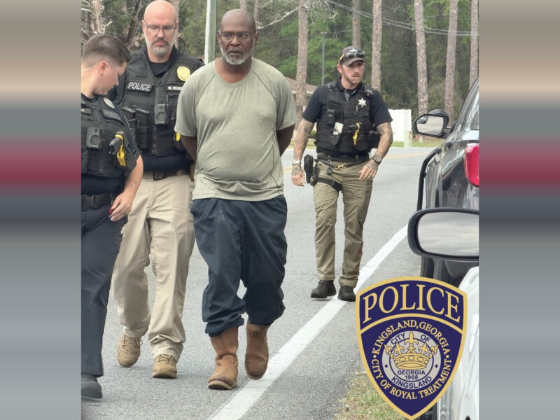 A man with a gray beard and glasses, wearing a light gray t-shirt, dark pants, and brown boots, is being escorted by three police officers on a road. The officers are in uniform with tactical vests, and one officer is walking behind the man while the other two are beside him. The scene takes place outdoors with trees in the background. A police badge for Kingsland, Georgia, is visible in the lower right corner.