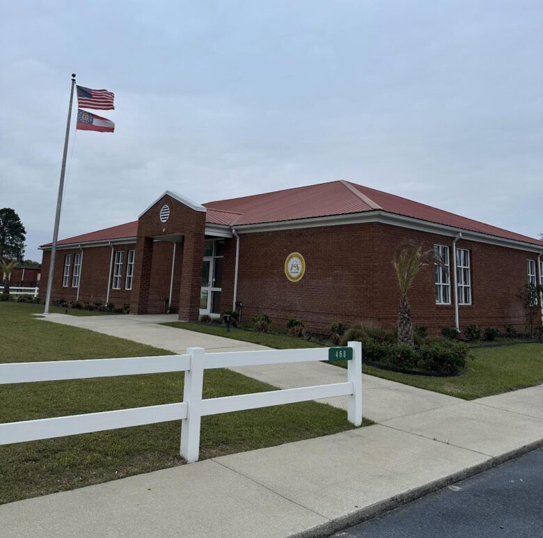 Single-story brick building with a red metal roof, multiple white-framed windows, and a covered entrance. Two flags, the American flag and the Georgia state flag, are flying on a flagpole near the entrance. A white fence with the number 468 is in the foreground, along a concrete sidewalk and green lawn with small shrubs and a palm tree. The sky is overcast.