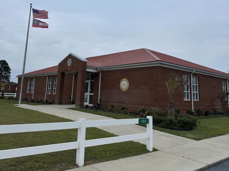 Single-story brick building with a red metal roof, multiple white-framed windows, and a covered entrance. Two flags, the American flag and the Georgia state flag, are flying on a flagpole near the entrance. A white fence with the number 468 is in the foreground, along a concrete sidewalk and green lawn with small shrubs and a palm tree. The sky is overcast.