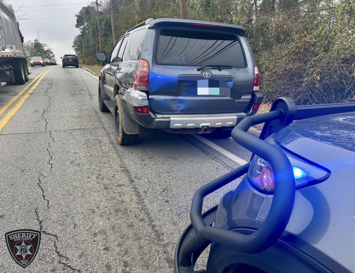 A dark-colored Toyota 4Runner with visible rear-end damage and taped rear left side is parked on the side of a road. A police vehicle with a push bumper and blue flashing lights is positioned behind it. The road has two yellow lines in the center, and there are other vehicles further ahead. Trees and bushes line the right side of the road. A sheriff's badge logo for Bibb County, Macon, is visible in the bottom left corner.