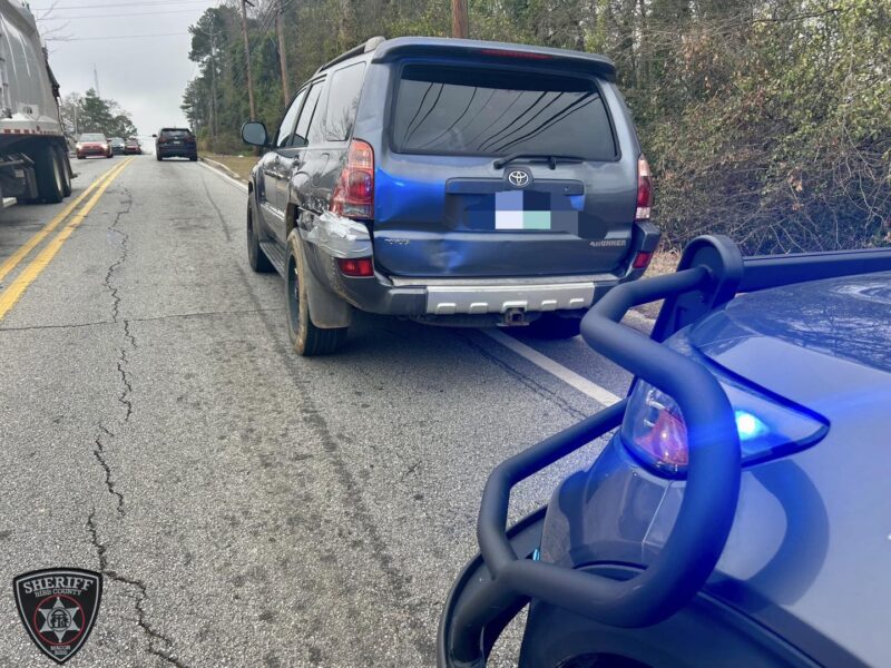 A dark-colored Toyota 4Runner with visible rear-end damage and taped rear left side is parked on the side of a road. A police vehicle with a push bumper and blue flashing lights is positioned behind it. The road has two yellow lines in the center, and there are other vehicles further ahead. Trees and bushes line the right side of the road. A sheriff's badge logo for Bibb County, Macon, is visible in the bottom left corner.