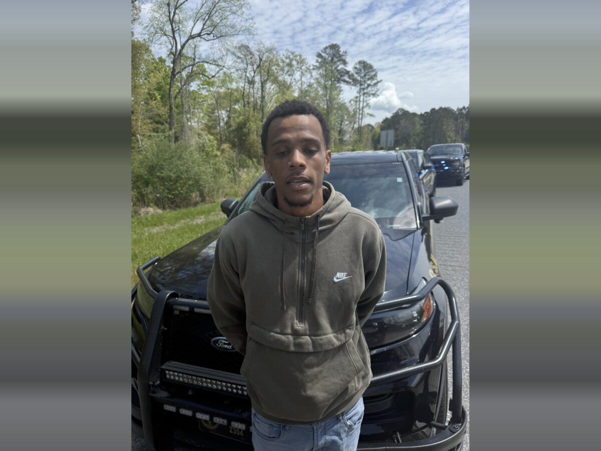 A young man wearing a green Nike hoodie and light blue jeans stands in front of a black Ford police SUV with a push bumper. The man has his hands behind his back, and there are additional police vehicles with flashing lights visible in the background on a road lined with trees and grass under a partly cloudy sky.