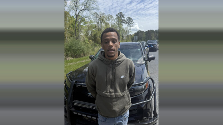 A young man wearing a green Nike hoodie and light blue jeans stands in front of a black Ford police SUV with a push bumper. The man has his hands behind his back, and there are additional police vehicles with flashing lights visible in the background on a road lined with trees and grass under a partly cloudy sky.