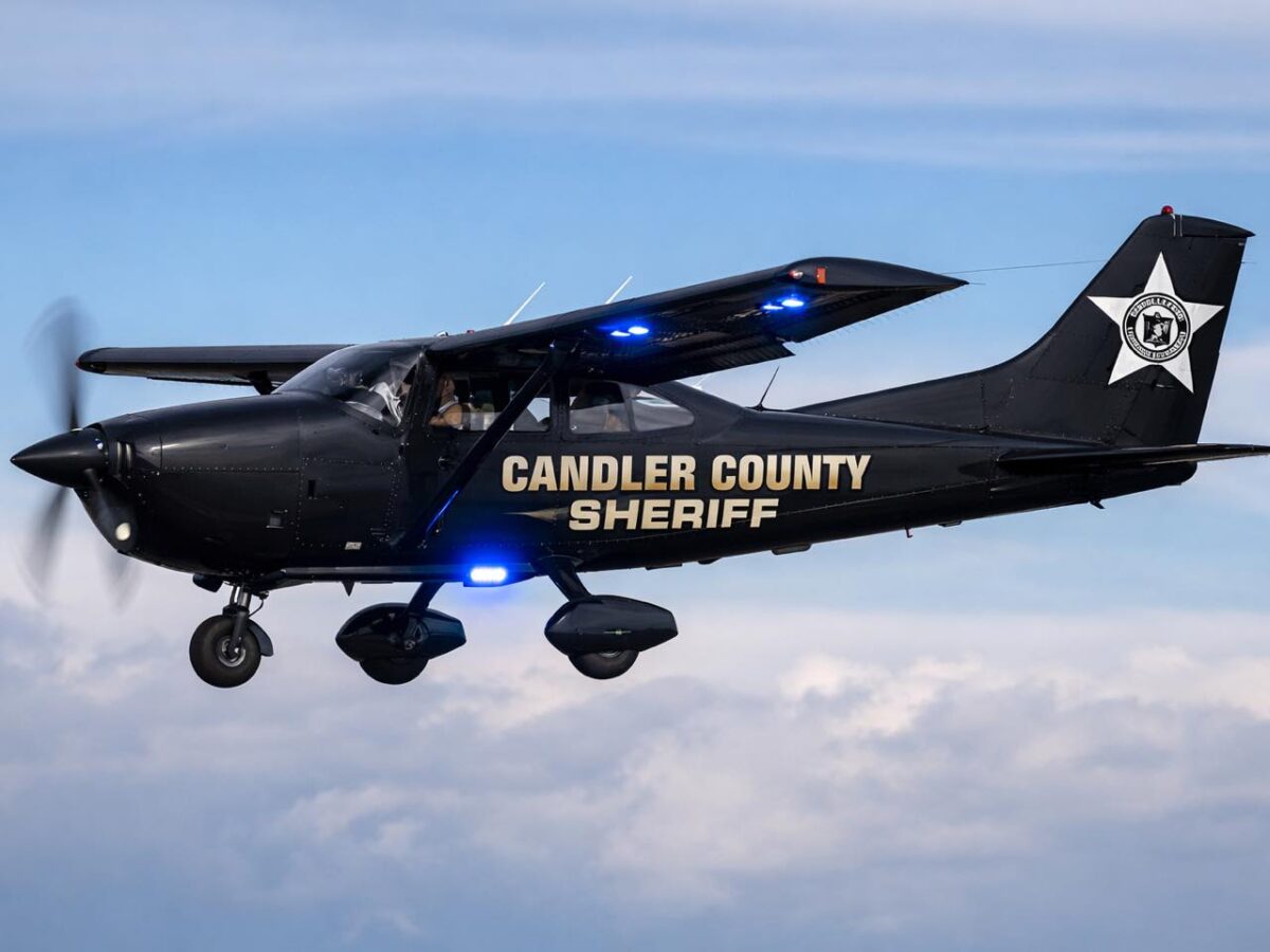 Black Candler County Sheriff airplane in flight with blue lights on the underside of the wings and landing gear, against a partly cloudy sky.