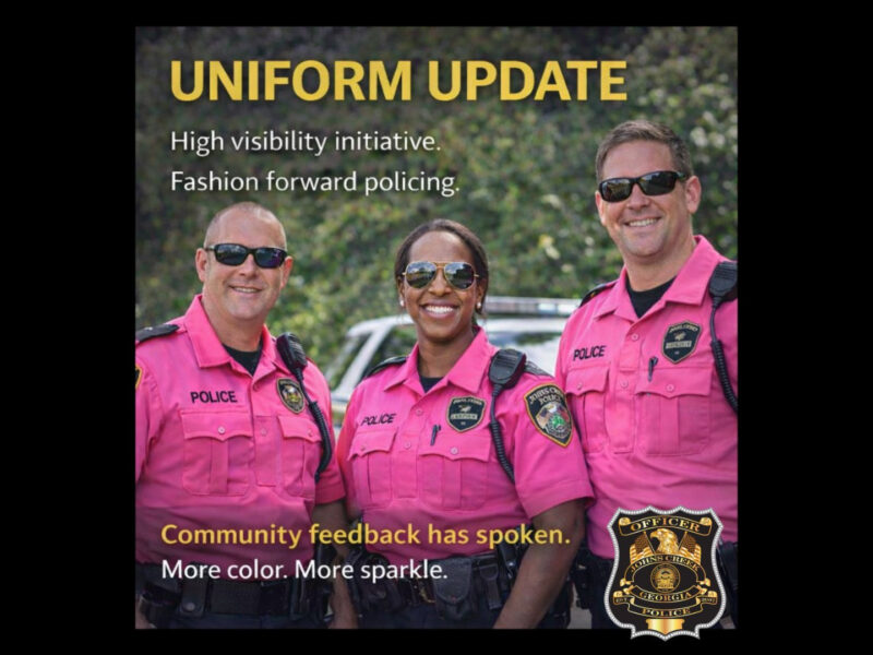 Three police officers wearing bright pink uniforms and sunglasses stand smiling in front of a police vehicle. The text above them reads "UNIFORM UPDATE" followed by "High visibility initiative. Fashion forward policing." Below the officers, the text says "Community feedback has spoken. More color. More sparkle." A police badge labeled "Johns Creek Police, Georgia" is displayed in the bottom right corner.