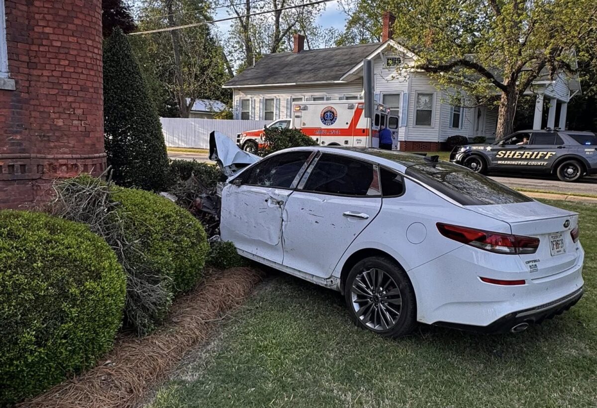 A white sedan with significant front-end damage is crashed into a brick building and bushes. The driver's side door is dented, and the hood is crumpled. In the background, there is an ambulance and a sheriff's vehicle parked near a white house with trees around.
