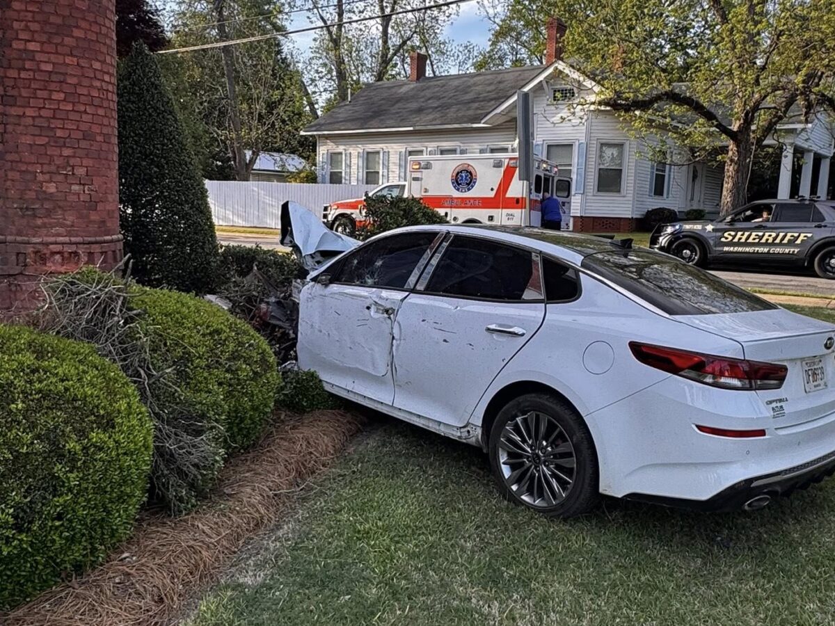 A white sedan with significant front-end damage is crashed into a brick building and bushes. The driver's side door is dented, and the hood is crumpled. In the background, there is an ambulance and a sheriff's vehicle parked near a white house with trees around.