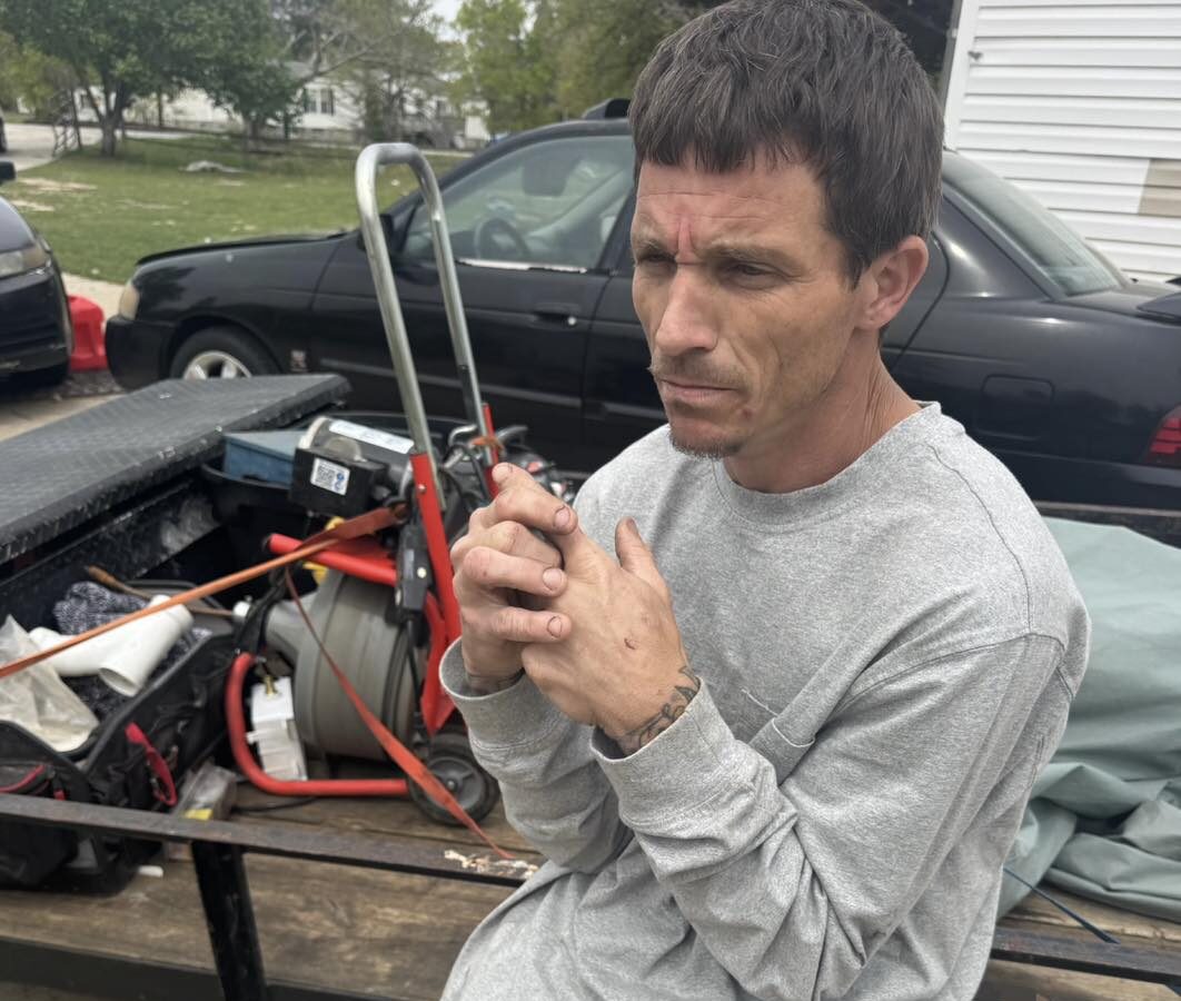 A man with short dark hair wearing a light gray long-sleeve shirt and tan cargo pants is sitting on the edge of a utility trailer. The trailer contains various tools and equipment, including a red dolly and a coiled hose. The man has a tattoo on his left wrist and appears to be clasping his hands together while looking to the side with a serious expression. A black car is parked behind the trailer, and there is a sheriff's badge logo in the bottom left corner of the image.